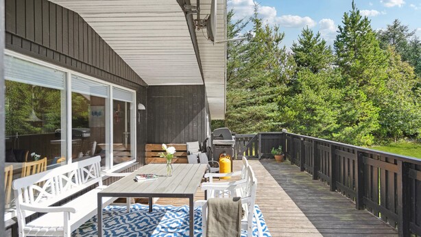 Una terraza de madera con mobiliario exterior, una parrilla y vistas a un área boscosa.