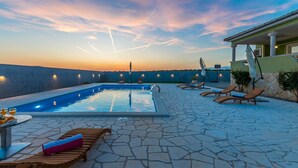 An outdoor swimming pool with lounge chairs and a table, surrounded by stone tiles and a sunset view. - Gorgeous home in Gornje Rastane (Gornje Rastane)