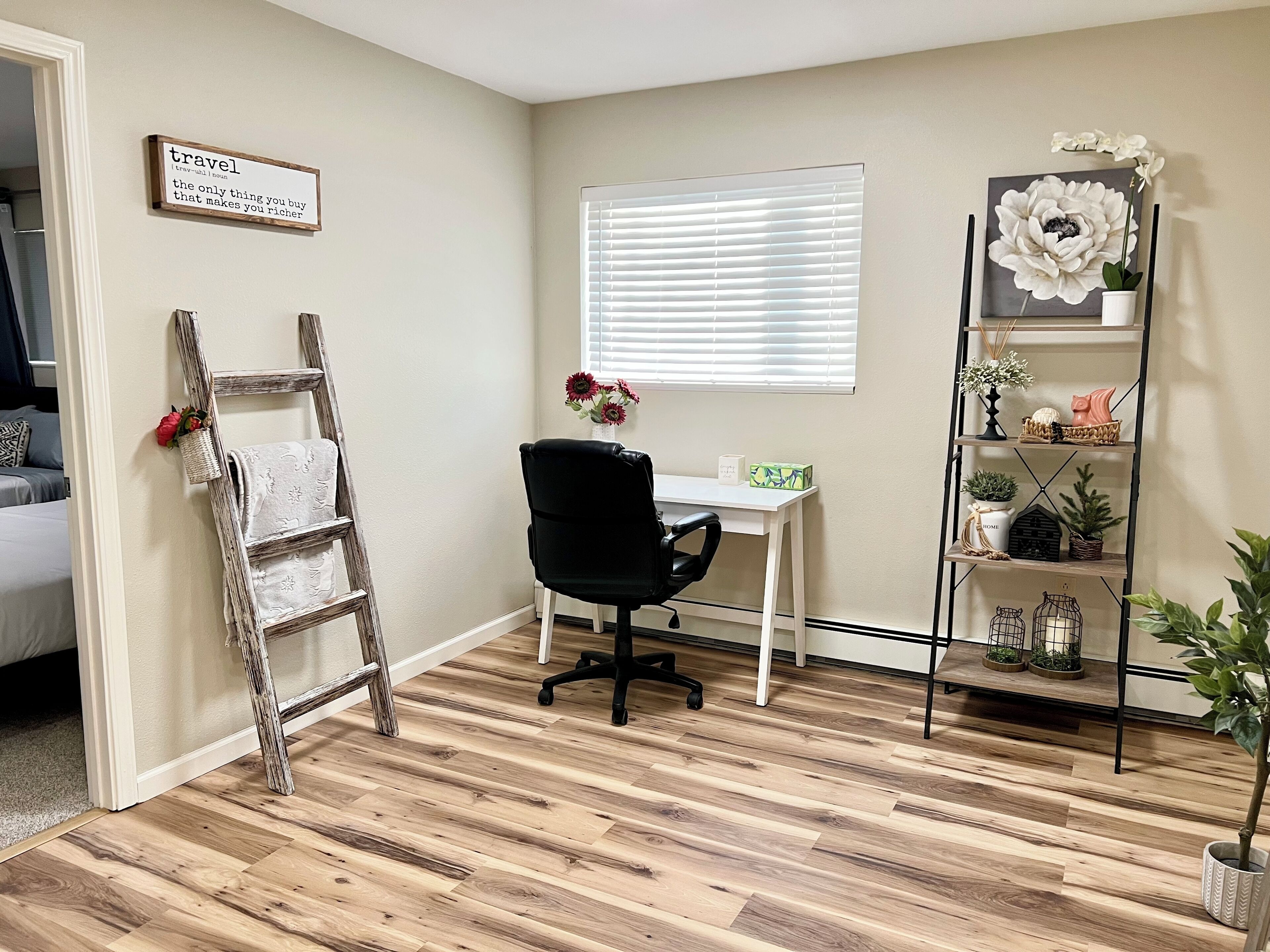 A room with a wooden floor, a white desk with a black office chair, a window with blinds, a ladder leaning against the wall, and a shelf with decorative items.
