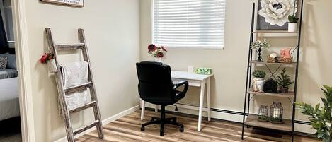 A room with a wooden floor, a white desk with a black office chair, a window with blinds, a ladder leaning against the wall, and a shelf with decorative items.