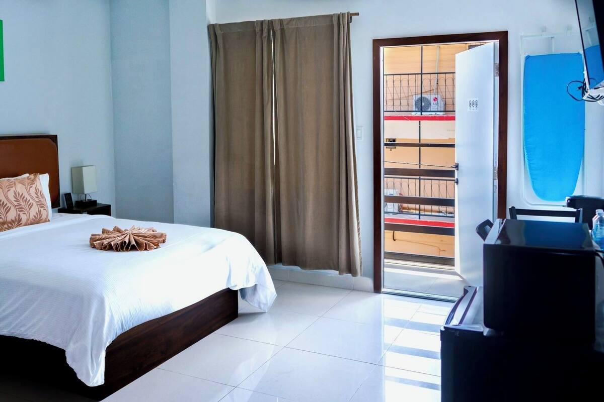 Ein Hotelzimmer mit Bett, Nachttisch, Lampe, Wandbild mit grünem Dekor, einem Fenster mit Vorhängen und Blick auf den Balkon.
