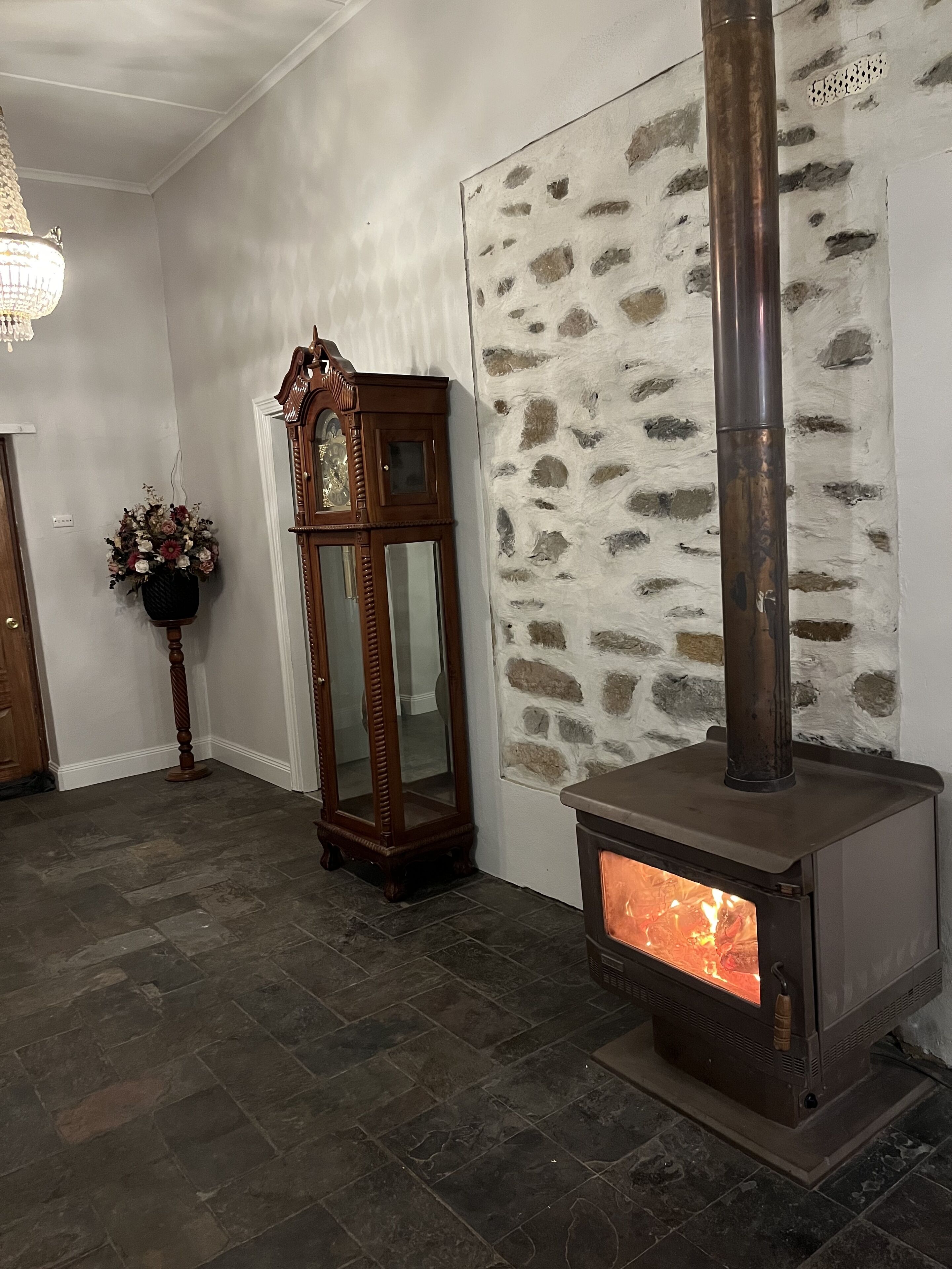 Une pièce avec un mur en pierre, une grande horloge à coucou en bois, un grand meuble en bois et un poêle à bois.