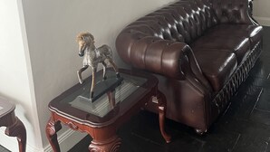 A living room with a brown leather sofa, a glass-top coffee table, and a coat rack with jackets. There are several framed pictures on the wall. - Magnificent historic homestead (Birdwood)