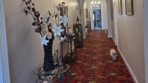 A hallway with a red patterned carpet, white walls, and a chandelier. There are framed pictures on the walls and a vase with flowers on a table. - Magnificent historic homestead (Birdwood)