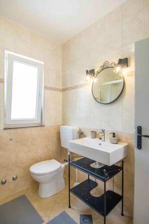A bathroom with a white toilet, a white sink with a black shelf, a round mirror with lights, and beige tiled walls. - Holiday apartment mit Terrasse (Biograd na Moru)