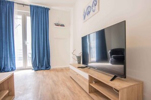 A modern living room with a large flat-screen TV on a wooden TV stand, blue curtains, and a sliding glass door leading to an outdoor area. - Holiday apartment mit Terrasse (Biograd na Moru)