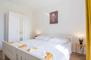 Uma cama bem arrumada com roupa de cama branca e toalhas amarelas, um guarda-roupa branco, uma mesa de cabeceira com abajur e uma obra de arte na parede.