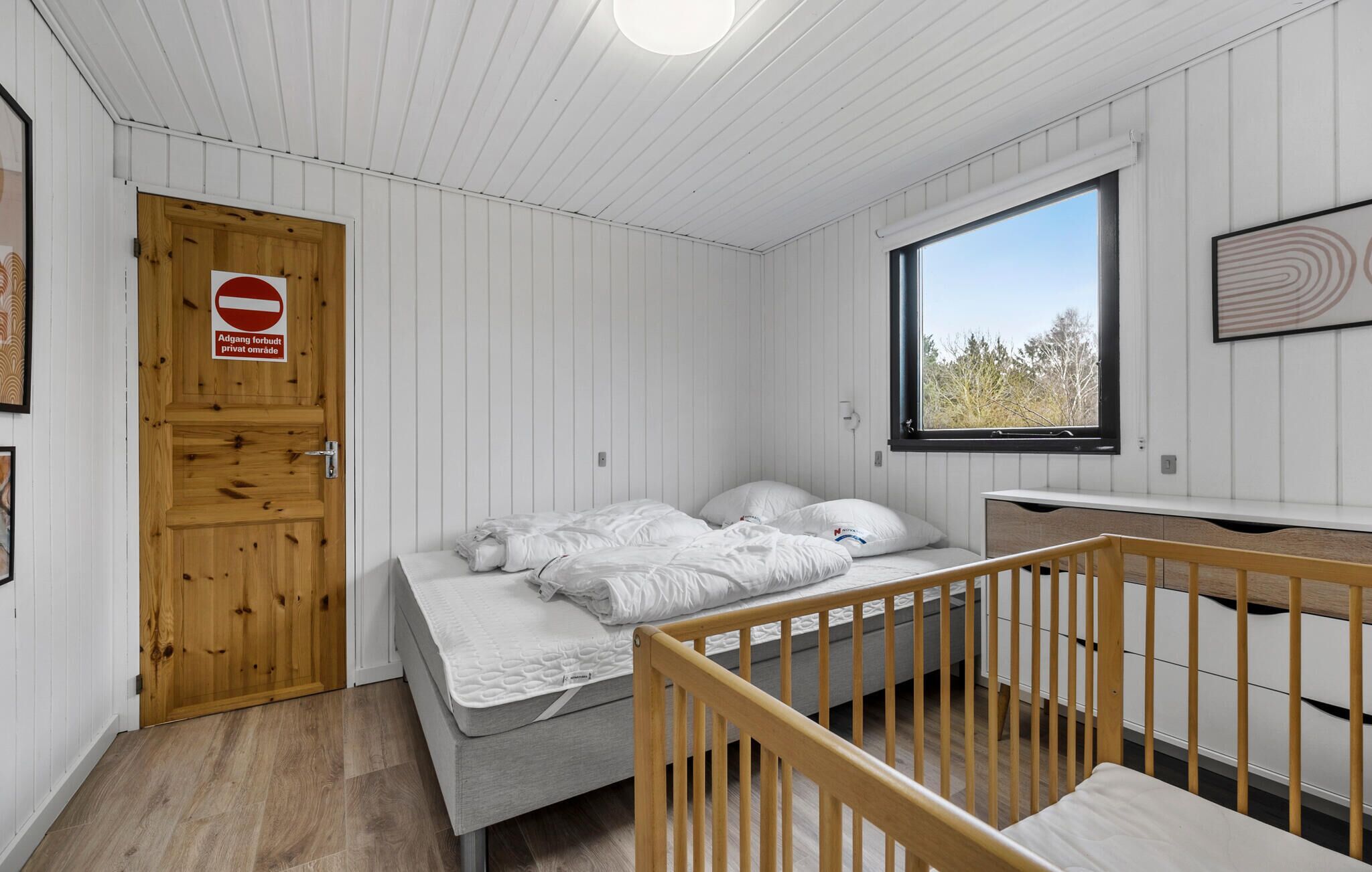 Une chambre à coucher avec un lit, un berceau, une fenêtre donnant sur un beau paysage, une porte avec une pancarte et un mur orné d’un tableau encadré.