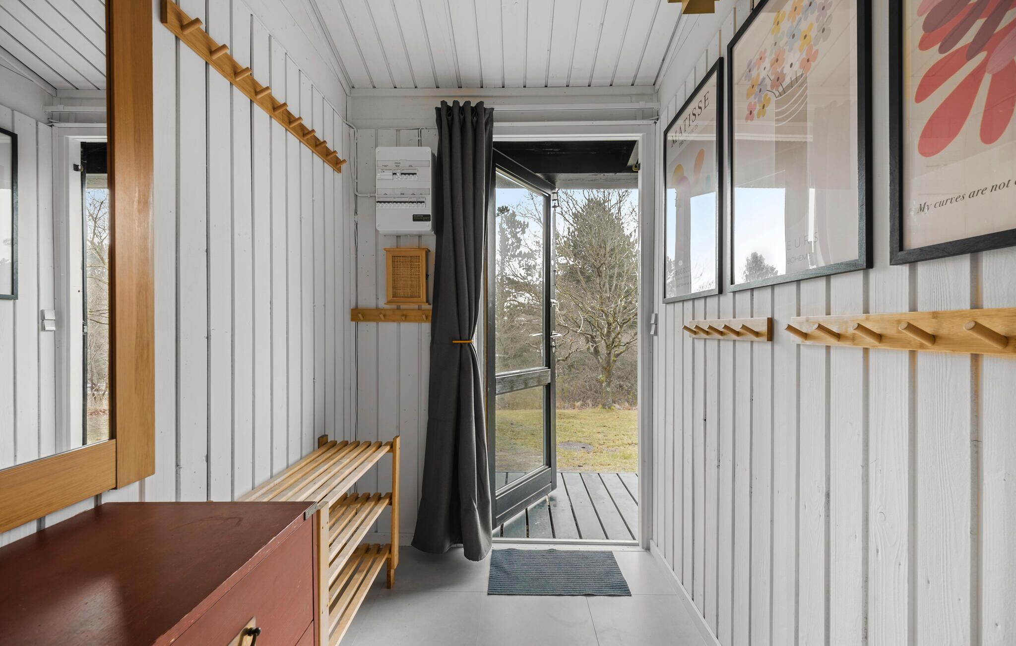 A narrow hallway with wooden walls and benches, leading to an open door with a view of trees.