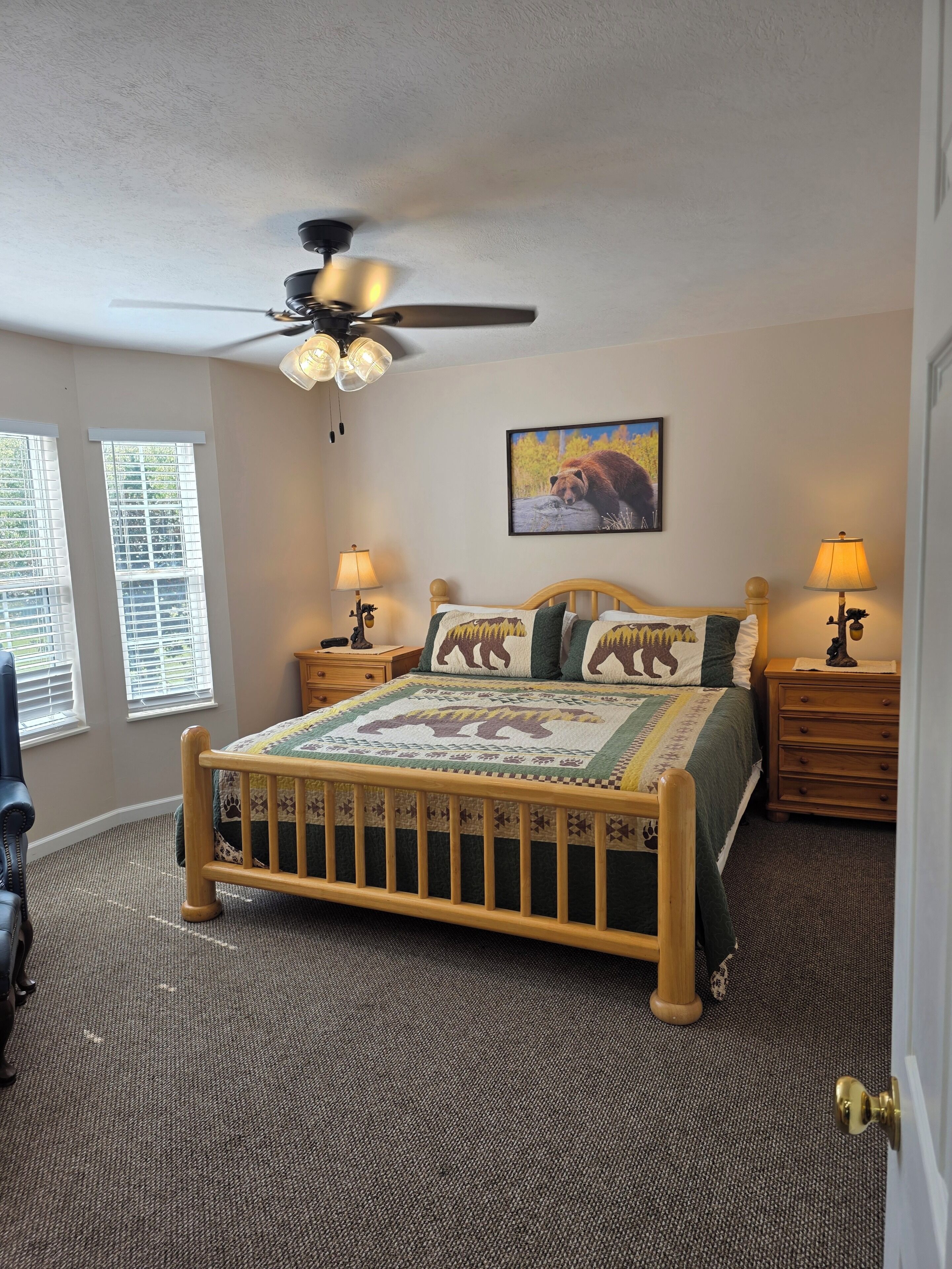 A bedroom with a wooden bed, two bedside tables with lamps, a ceiling fan, a window with blinds, and a wall-mounted picture.