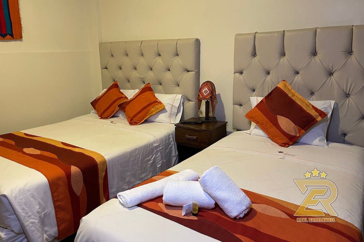 A hotel room with two beds, each with a tufted headboard, white bedding, and orange and brown patterned pillows. There is a nightstand with a lamp and a decorative item between the beds.