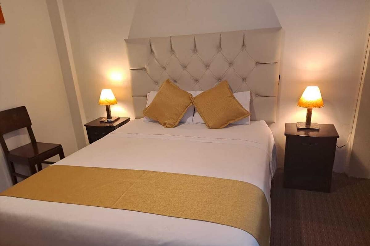 A hotel room with a large bed, two bedside tables with lamps, a wooden chair, and a headboard.
