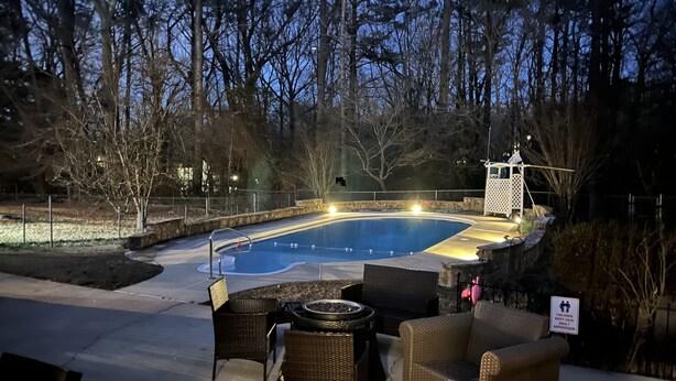 An outdoor swimming pool with lounge chairs and a fire pit, surrounded by trees and a stone wall.