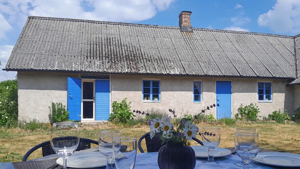 A rustic house with a thatched roof, blue shutters, and a dining table set outdoors with wine glasses and a vase of flowers. - 2 bedroom cozy home in Munka Ljungby (Munka Ljungby)