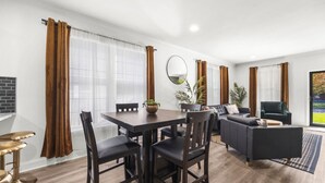 A modern living room with a dark wood dining table, black chairs, a grey sofa, and a dark armchair. There is a large window with brown curtains, a mirror on the wall, and a potted plant on the table.