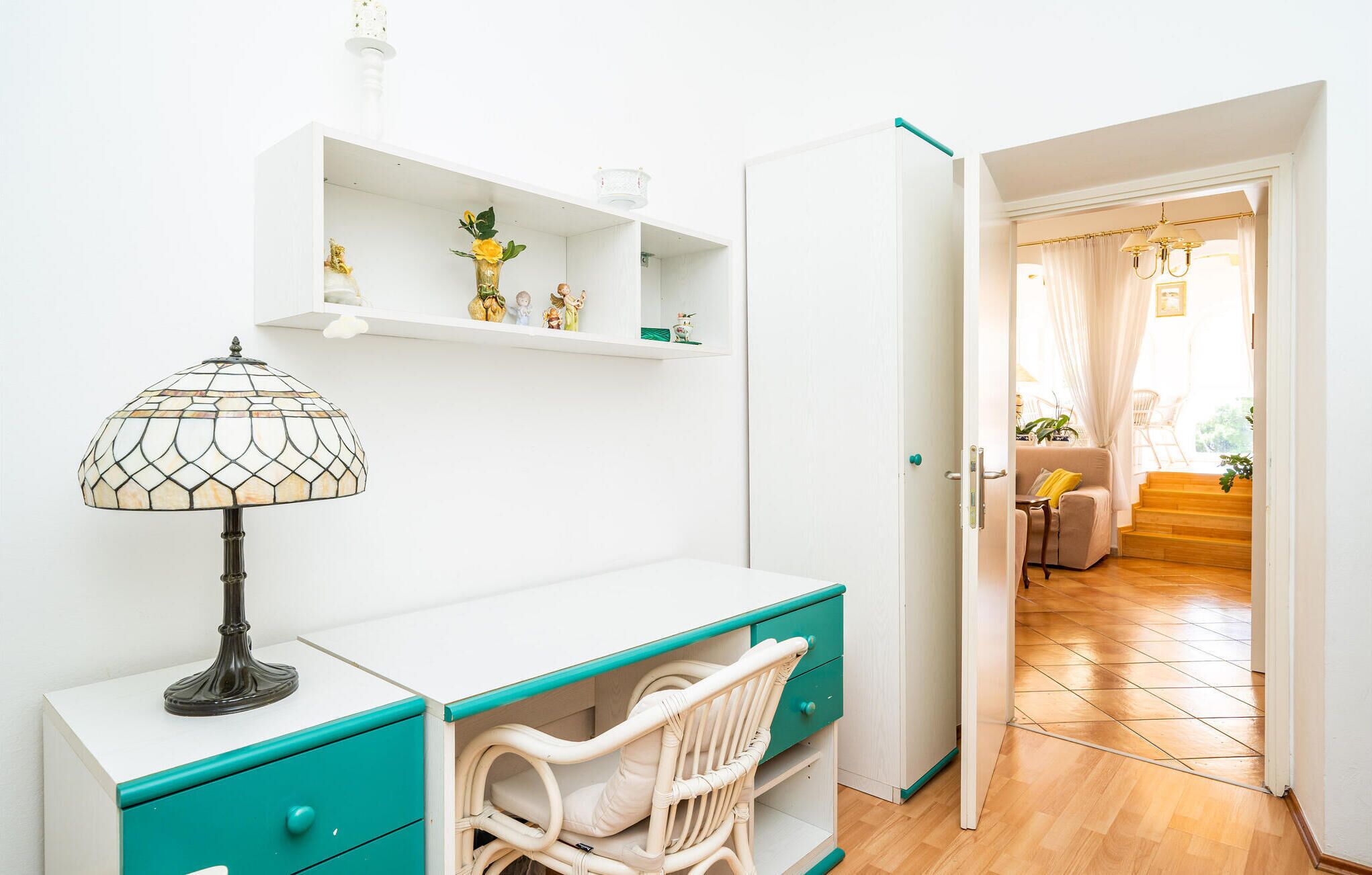 Une pièce avec un bureau blanc, un tiroir turquoise, une chaise en osier et une lampe. On y trouve également une armoire blanche et une porte donnant sur une autre pièce.