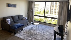 A living room with a grey sofa, a small round table, a large window with curtains, and a sign on the wall.