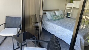 A balcony with a glass door, two chairs, a small table, a bed with a blue bedspread and pillows, and a nightstand with a lamp.