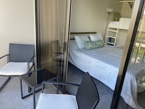 A balcony with a glass door, two chairs, a small table, a bed with a blue bedspread and pillows, and a nightstand with a lamp.
