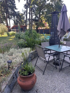 An outdoor patio area with a table and chairs, surrounded by a garden with various plants and a large terracotta pot. - Garden-Level Suite in Quiet Colwood Neighbourhood (Victoria)