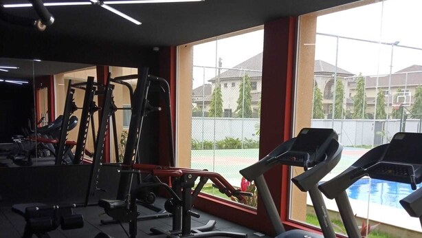 A gym with treadmills, weight machines, and a large window with a view of a swimming pool and buildings. - Modern 2 Bedroom Duplex - Gated Estate , 24/7 Security, Gym, Pool, Tennis Court (Tema)