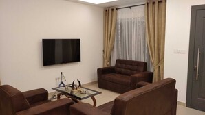 A living room with a flat-screen TV mounted on the wall, a brown leather sofa, a glass coffee table, and beige curtains. - Modern 2 Bedroom Duplex - Gated Estate , 24/7 Security, Gym, Pool, Tennis Court (Tema)