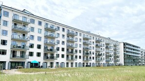 A multi-story residential building with balconies, a sandy foreground, and a clear sky.