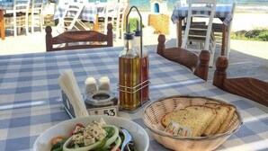 A table set with a bottle of olive oil, a bowl of salad, and a basket of bread on a beach with a view of the sea. - Liopetri 2-Bed Apt, Sleeps 6, Pool, 10min to Beach (Liopetri)