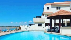 A swimming pool with lounge chairs and a gazebo, surrounded by a building with a tiled roof. - Liopetri 2-Bed Apt, Sleeps 6, Pool, 10min to Beach (Liopetri)