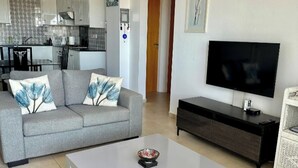 A living room with a grey sofa, a white coffee table, a black TV stand, and a wicker chair. - Liopetri 2-Bed Apt, Sleeps 6, Pool, 10min to Beach (Liopetri)