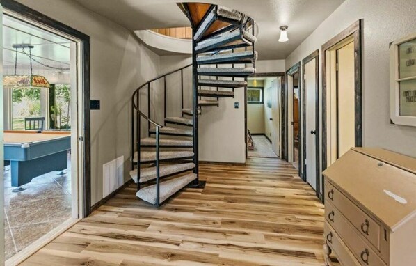 Un escalier en colimaçon dans un couloir au plancher en bois, une table de billard visible à travers une porte ouverte, et une commode sur la droite.