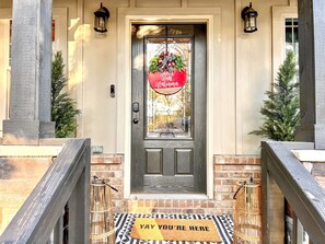 Una puerta principal con un cartel que dice 'Welcome Home Alabama', un felpudo con la inscripción 'YAY YOU'RE HERE' y una pequeña jaula de pájaros en los escalones.