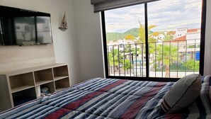 Ein Schlafzimmer mit einem großen Bett, einem an der Wand befestigten Fernseher und Blick auf eine Wohngegend durch ein Fenster.