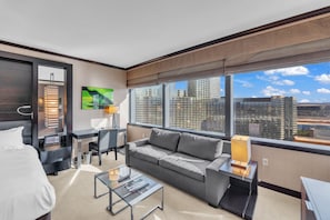 A modern hotel room with a large bed, a grey sofa, a glass coffee table, a desk with a chair, and a large window offering a city view.