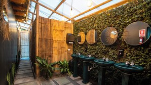 Un sendero estrecho que conduce a una puerta de madera, flanqueado por plantas verdes y tres lavamanos verdes con vasos.