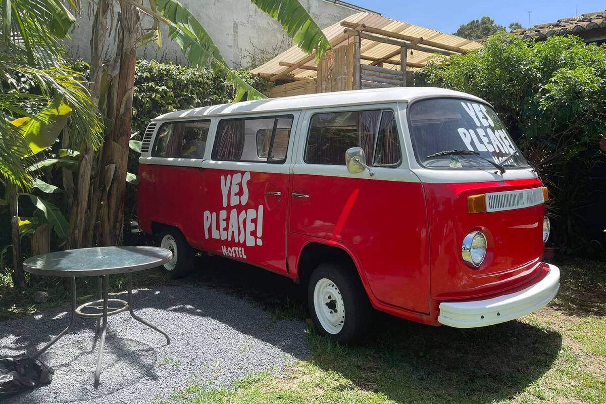 A red and white van with "YES PLEASE!" and "HOTEL" written on the side, parked in a garden with a table and chairs nearby.