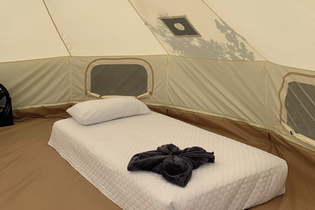 A single bed with a quilted white mattress, a black towel, and a white pillow inside a tent.