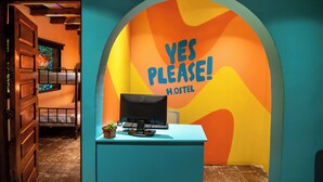 Una recepción con una computadora y una planta pequeña, una cama litera al fondo, y una pared colorida con el texto "YES PLEASE! H.OSTEL".