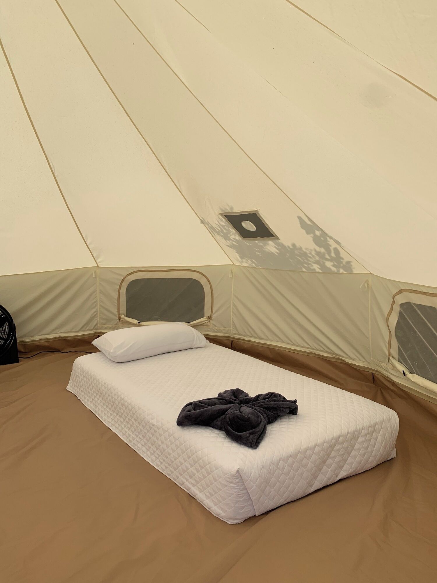 A single bed with a quilted white mattress, a black towel, and a white pillow inside a tent.