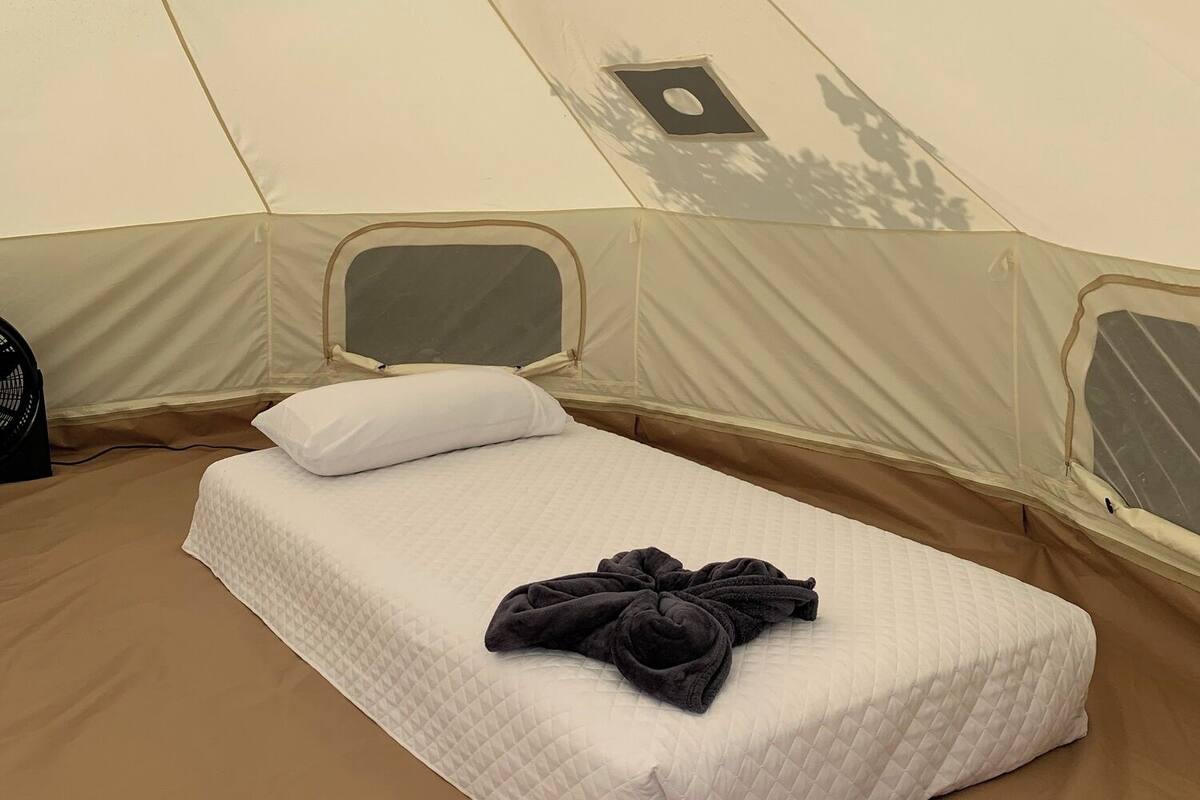 A single bed with a quilted white mattress, a black towel, and a white pillow inside a tent.