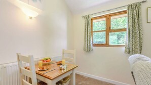 Un coin repas avec une table en bois, des chaises blanches, un panier de fruits et une fenêtre agrémentée de rideaux à motifs floraux.