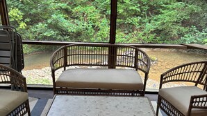 A wooden bench with a cushioned seat and backrest, a small table, and a striped mat on a balcony overlooking a forested area.