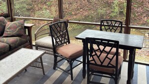 A covered outdoor seating area with wicker furniture, a table, and a view of a forested area.