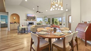 A spacious dining area with a wooden table set for six, surrounded by chairs. A modern chandelier hangs above, and a television is mounted on the wall.