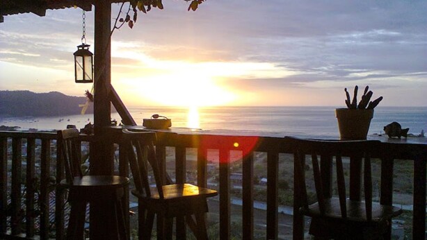 A wooden deck with chairs and a table, overlooking a sunset over the ocean.