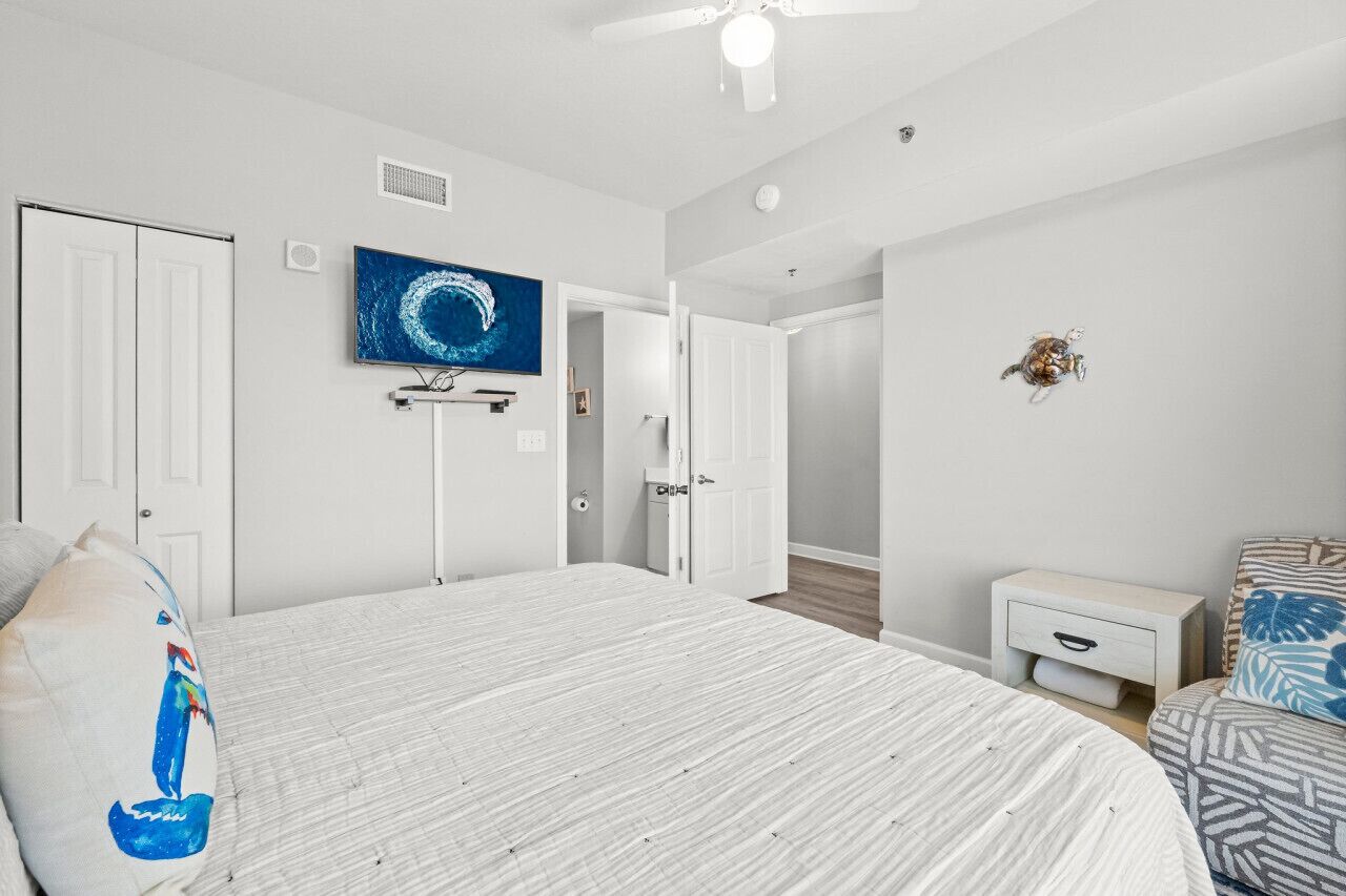 Un dormitorio con una cama grande, un ventilador de techo, un televisor en la pared y una mesita de noche pequeña.
