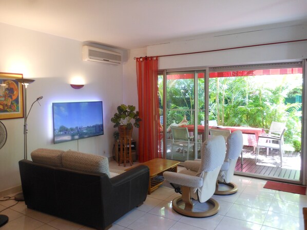 A living room with a sofa, armchairs, a TV, and a sliding glass door leading to an outdoor area with a table and chairs.