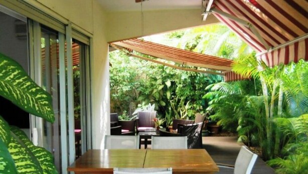 Une terrasse extérieure avec une table et des chaises en bois, entourée d’une végétation luxuriante et agrémentée d’un auvent à rayures.