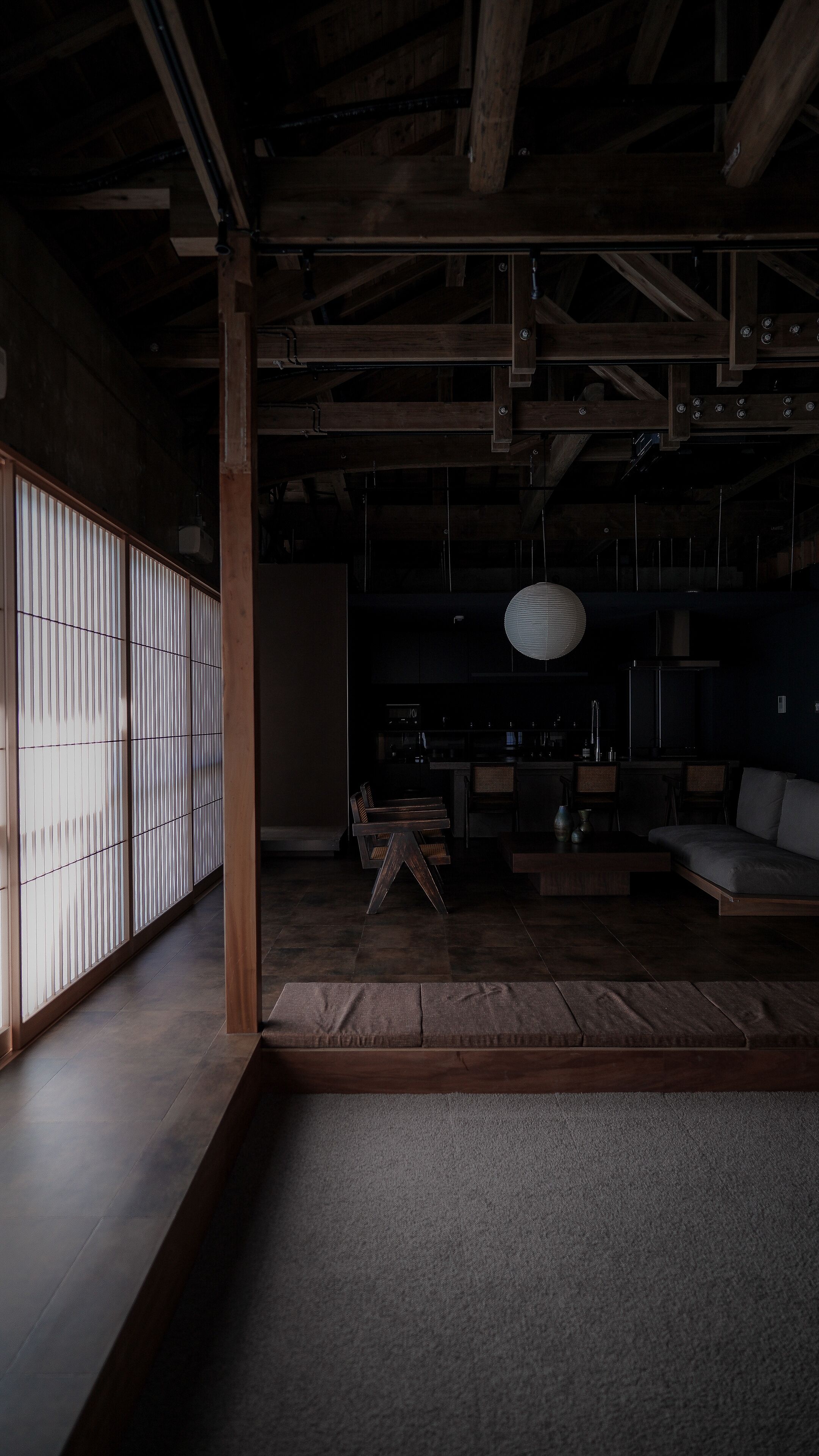 A dimly lit interior with a wooden ceiling, a traditional Japanese sliding door, a dining area with a low table and chairs, and a hanging paper lantern.