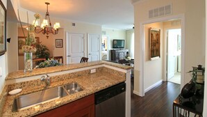 A modern kitchen with granite countertops, a stainless steel sink, and a dishwasher. There is a dining area with a table and chairs, and a chandelier hanging above. - The Palm Tree Penthouse (Stuart)